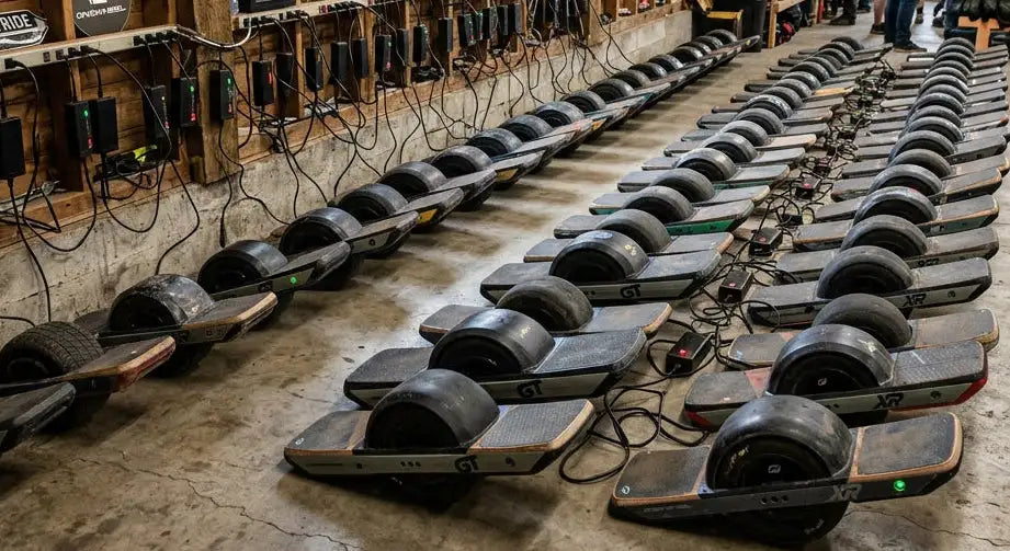 Rows of black and gray onewheel electric scooters with rubber tires are lined up in a warehouse.
